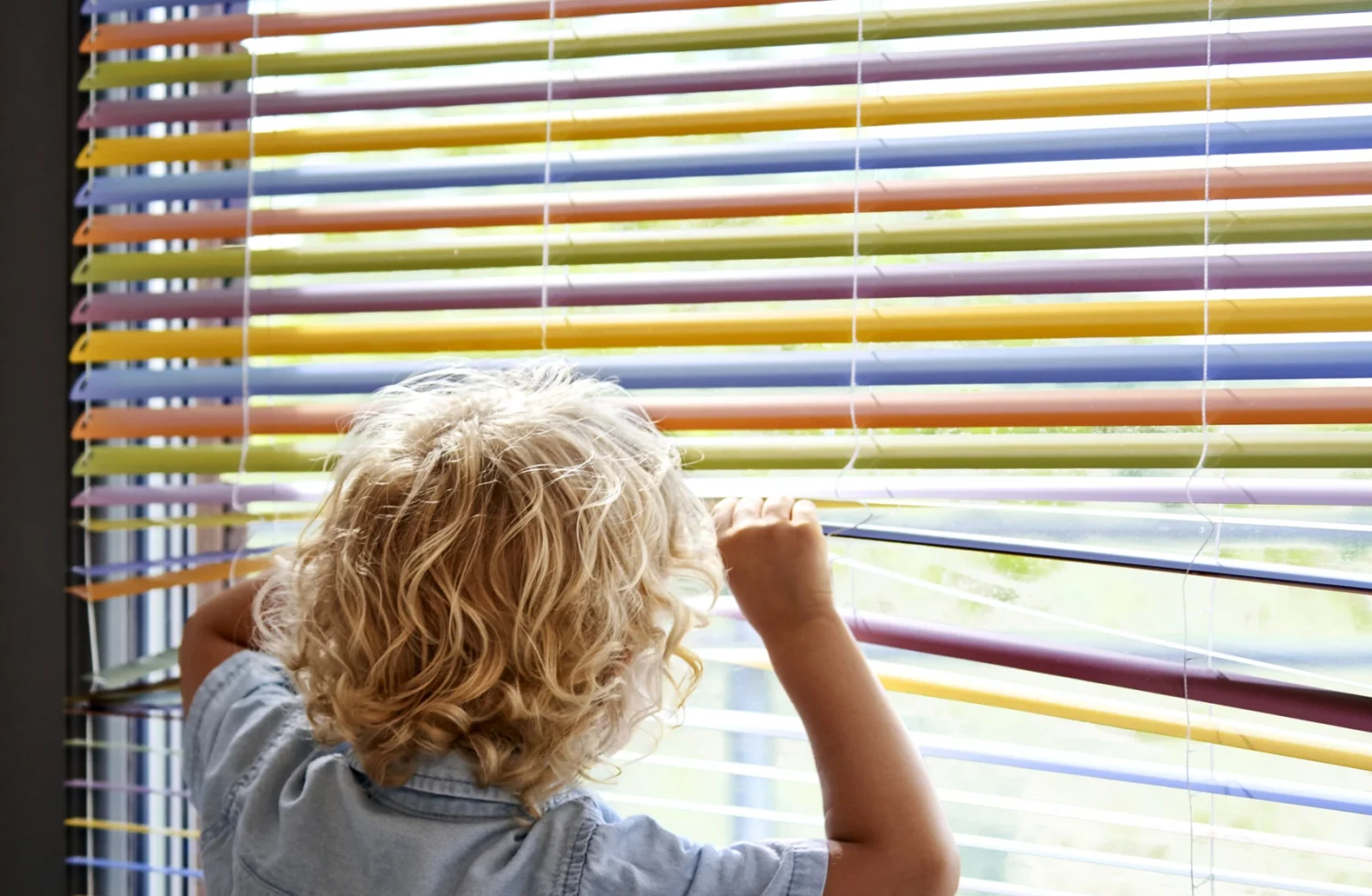 Kind schaut durch bunte Jalousien ans Fenster
